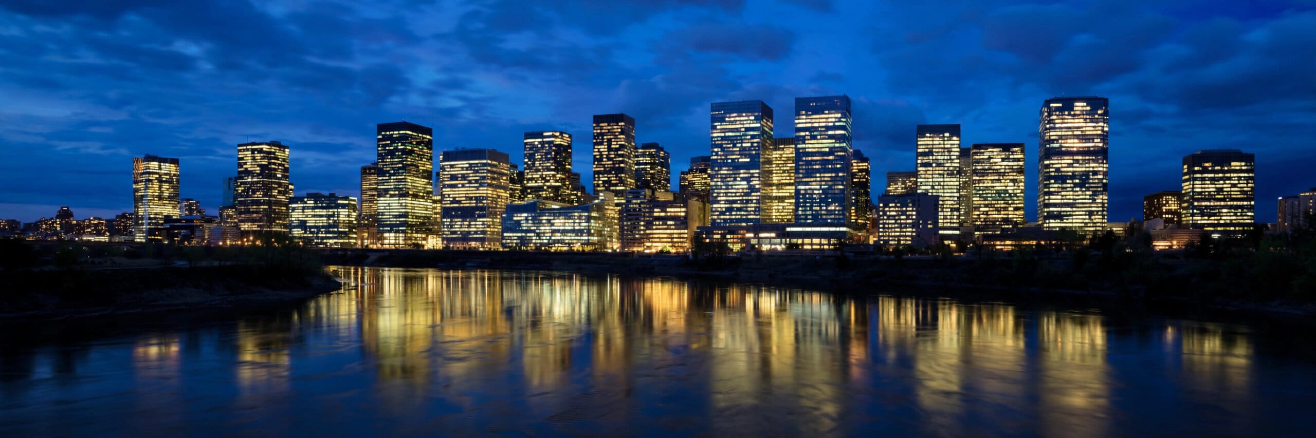 A financial district skyline with lit office towers reflected in a river at dusk